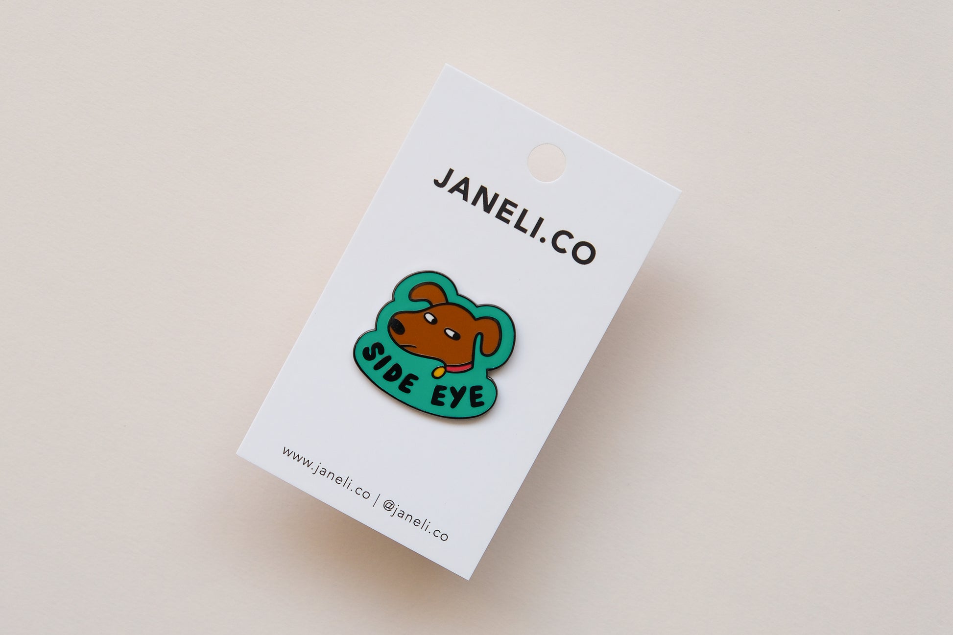 An enamel pin showing a sassy dog giving side eye that says "Side Eye" on a white JaneLi.Co backing card over a cream background.