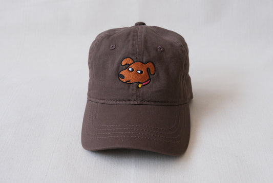 A photo of a chocolate brown dad hat with an embroidered dog giving side eye over a cream background.