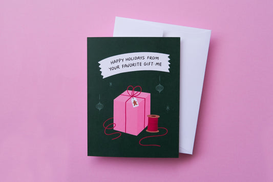 A photo of a greeting card that says "Happy Holidays From Your Favorite Gift: Me" with a present and a white envelope on a pink background.