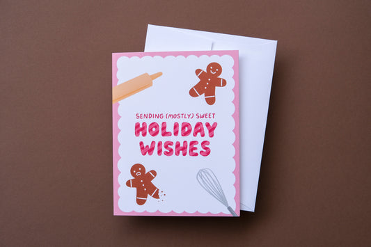 A photo of a greeting card that says "Sending (mostly) sweet holiday wishes" with two gingerbread cookies, one with a leg bitten off and a white envelope on a brown background.