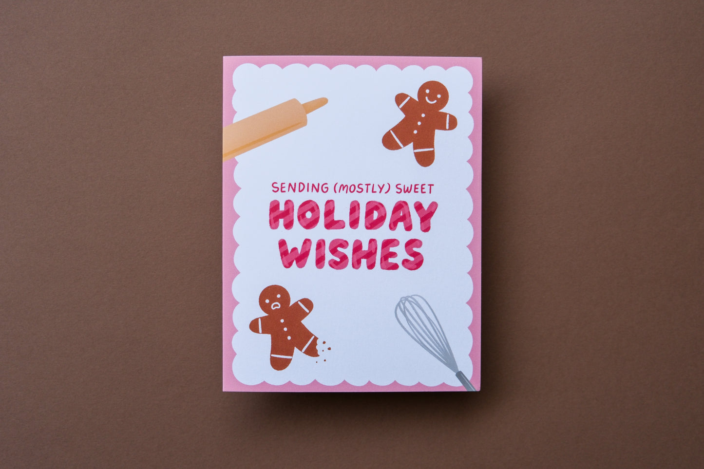 A photo of a greeting card that says "Sending (mostly) sweet holiday wishes" with two gingerbread cookies, one with a leg bitten off on a brown background.