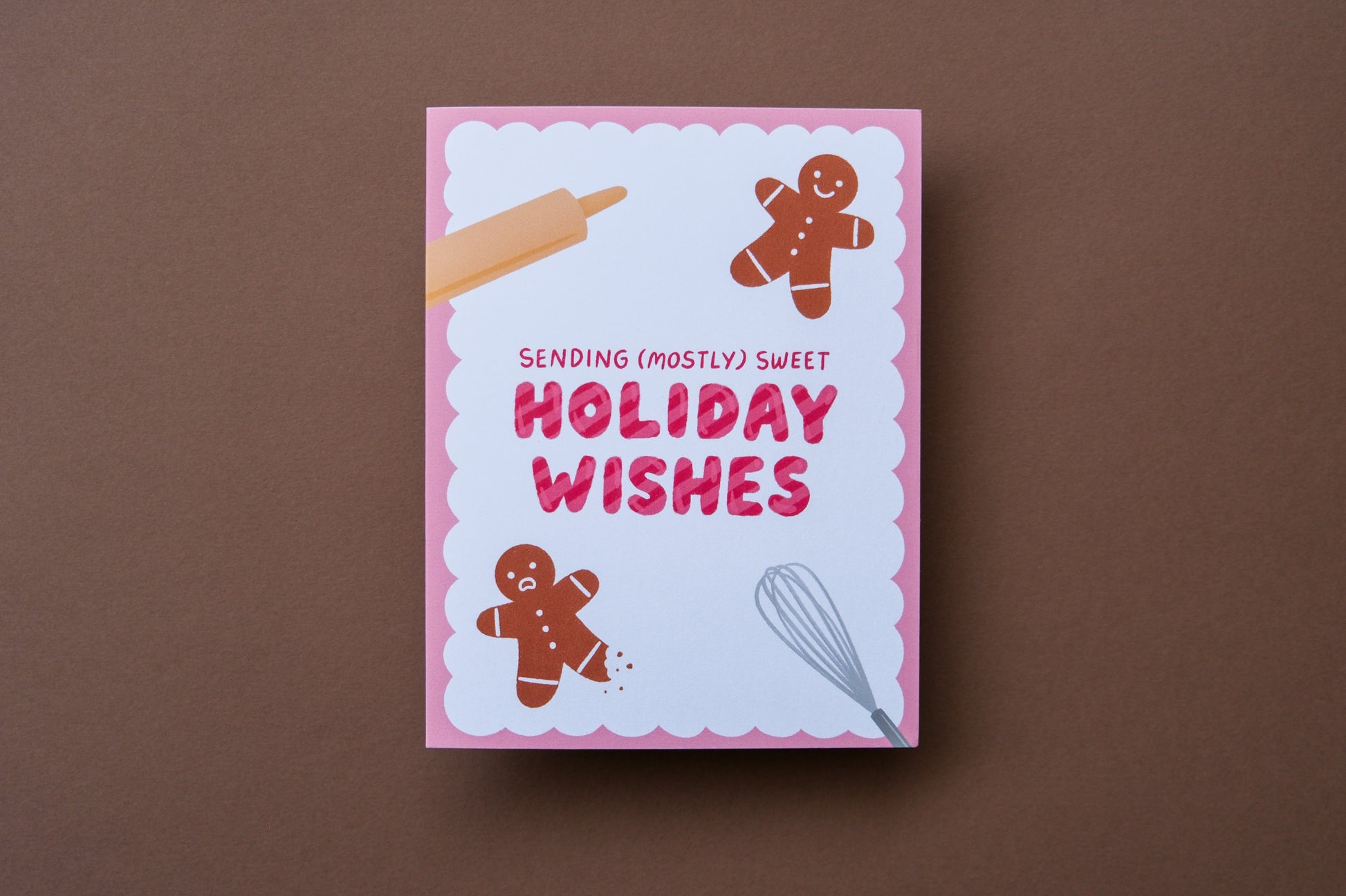 A photo of a greeting card that says "Sending (mostly) sweet holiday wishes" with two gingerbread cookies, one with a leg bitten off on a brown background.