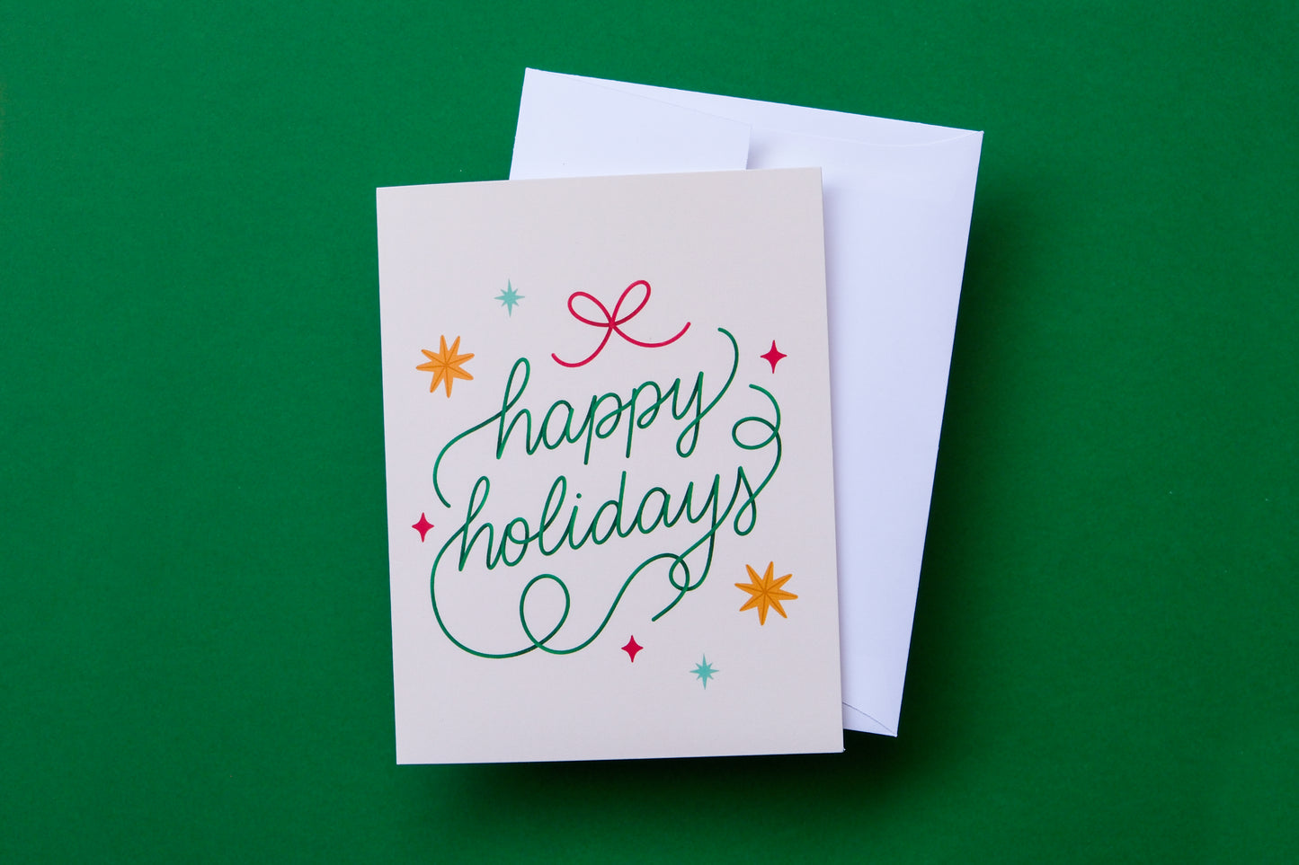 A photo of a greeting card that says "happy holidays" with stars and a bow and a white envelope on a green background.