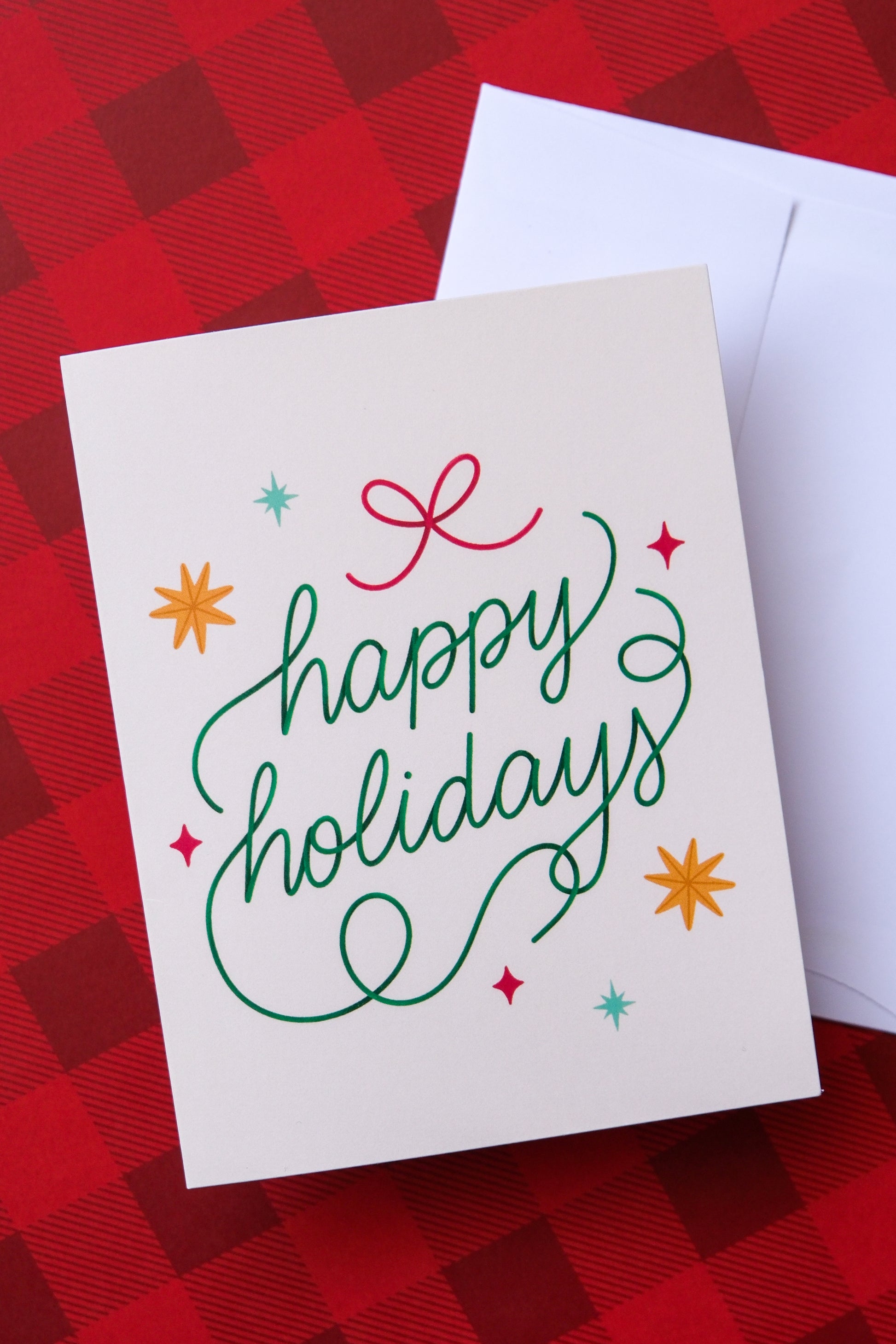 A photo of a greeting card that says "happy holidays" with stars and a bow and a white envelope on a red plaid background.