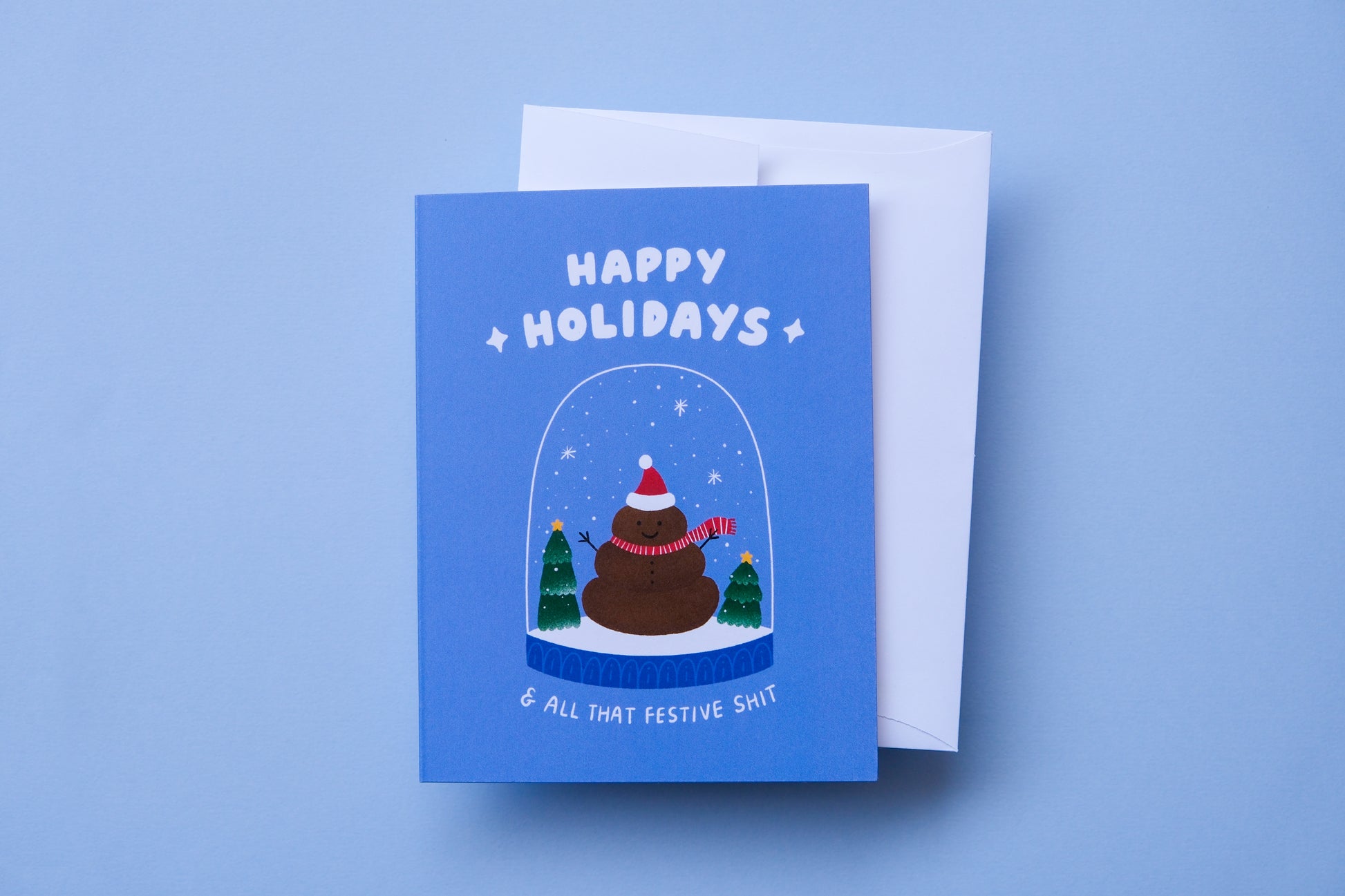 A photo of a greeting card that says "Happy holidays and all that festive shit" with a poop snowman in a snowglobe and a white envelope on a blue background.