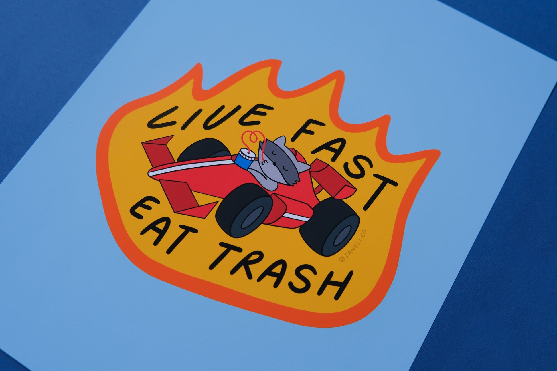 A close up of a JaneLi.Co print of a racoon driving a racecar while sipping a soda that says "Live Fast Eat Trash" over a blue background.