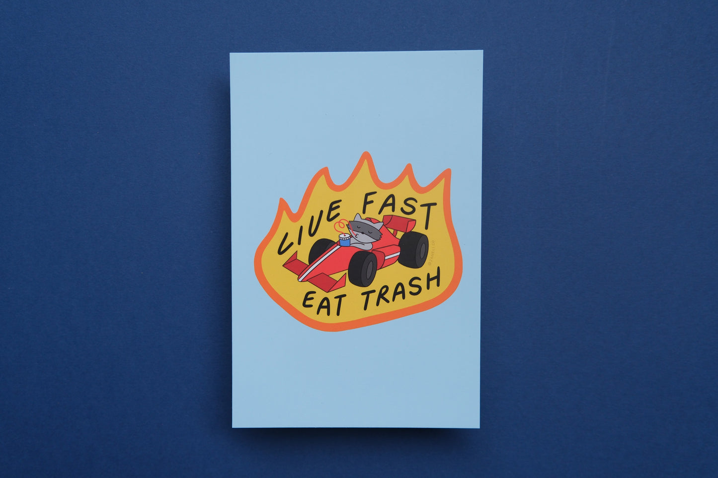 A JaneLi.Co mini print/postcard of a racoon driving a racecar while sipping a soda that says "Live Fast Eat Trash" over a blue background.