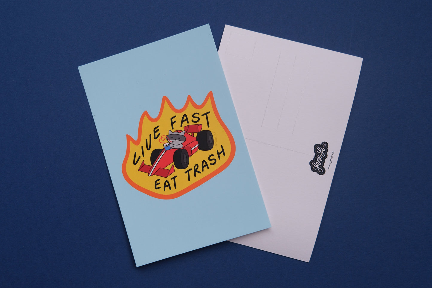 A JaneLi.Co mini print/postcard of a racoon driving a racecar while sipping a soda that says "Live Fast Eat Trash" and a back postcard side of the same postcard over a blue background.