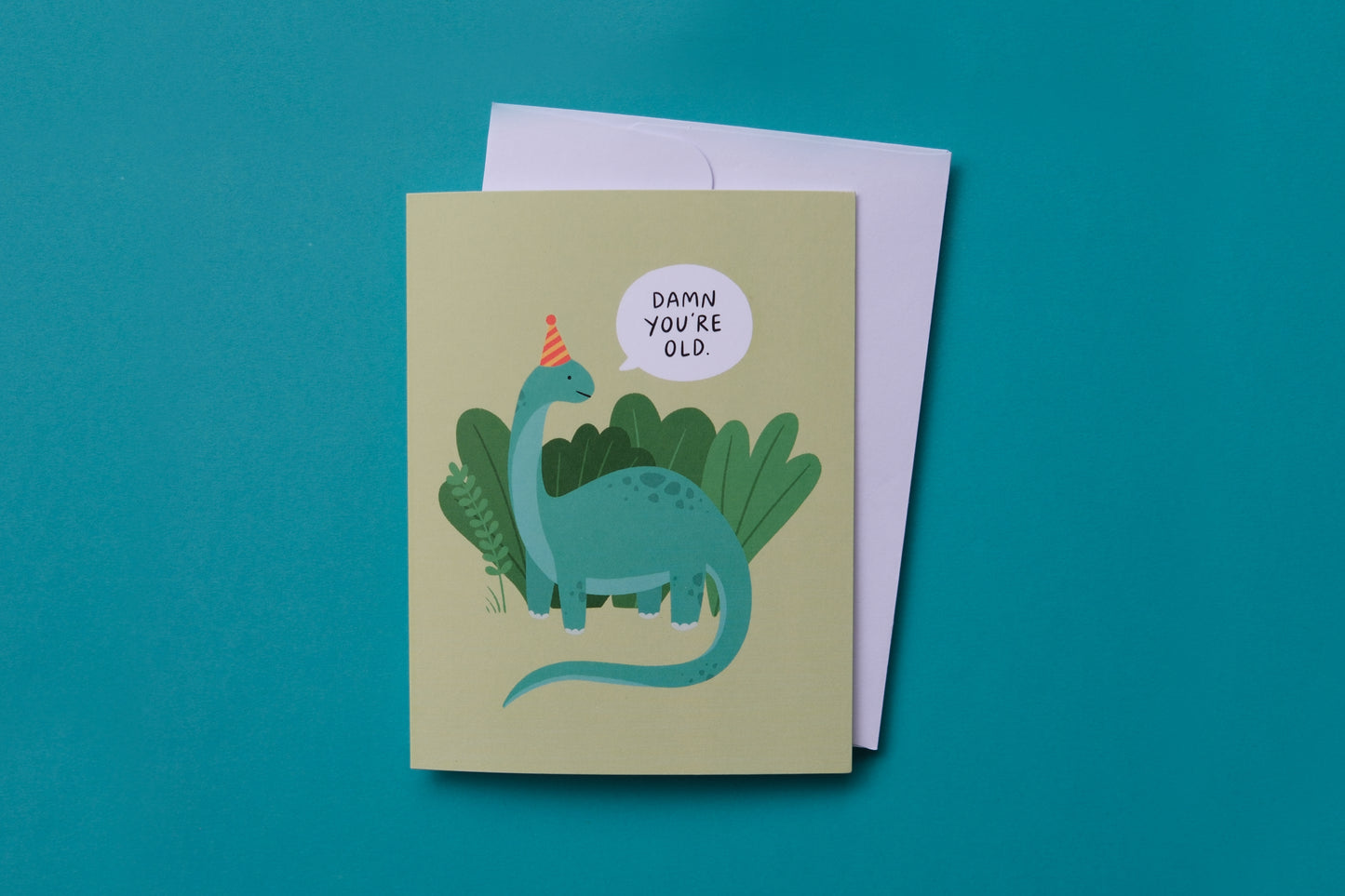 A photo of a greeting card with dinosaur saying "Damn you're old" and a white envelope on a turquoise background.