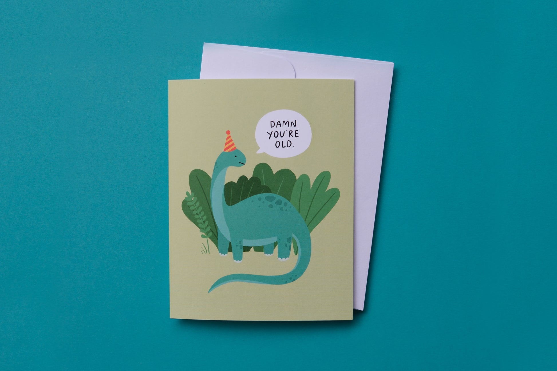 A photo of a greeting card with dinosaur saying "Damn you're old" and a white envelope on a turquoise background.