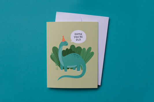 A photo of a greeting card with dinosaur saying "Damn you're old" and a white envelope on a turquoise background.