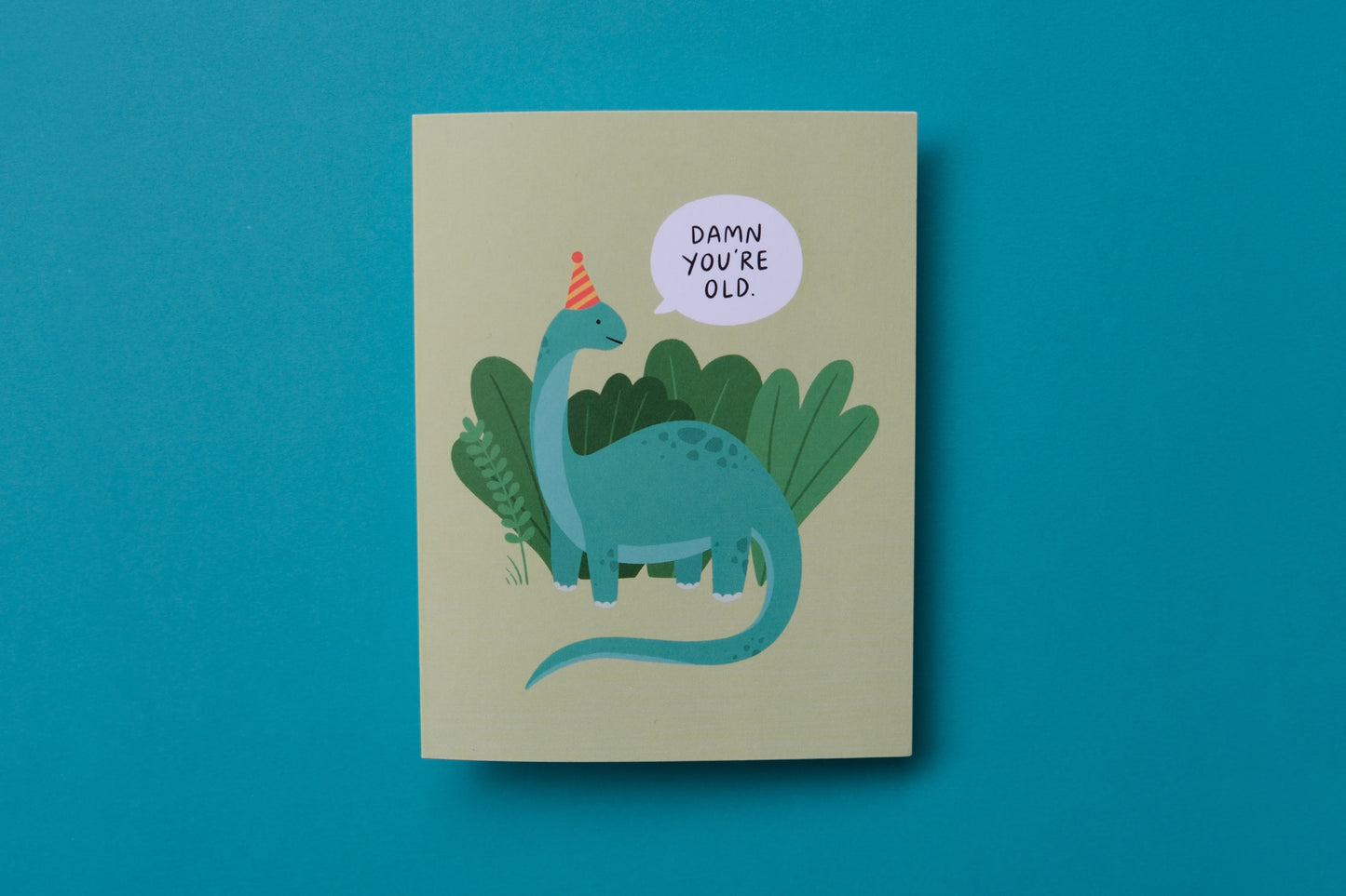 A photo of a greeting card with dinosaur saying "Damn you're old" on a turquoise background.