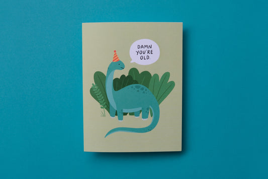 A photo of a greeting card with dinosaur saying "Damn you're old" on a turquoise background.
