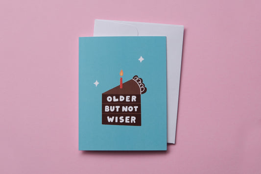 A photo of a greeting card that says "Older but not wiser" on a slice of chocolate cake and a white envelope on a pink background.