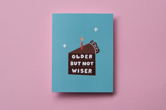 A photo of a greeting card that says "Older but not wiser" on a slice of chocolate cake on a pink background.