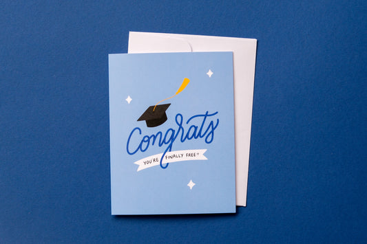 A photo of a greeting card that says "Congrats you're finally free!!" with a graduation cap and a white envelope on a dark blue background.