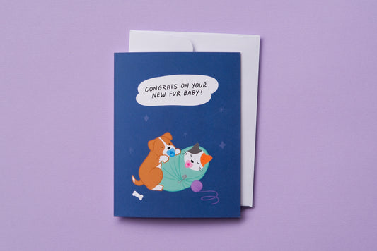 A photo of a greeting card that says "Congrats on your new fur baby!" with a puppy and a kitten swaddled and sucking on pacifiers and a white envelope on a lavender background.