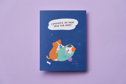 A photo of a greeting card that says "Congrats on your new fur baby!" with a puppy and a kitten swaddled and sucking on pacifiers on a lavender background.