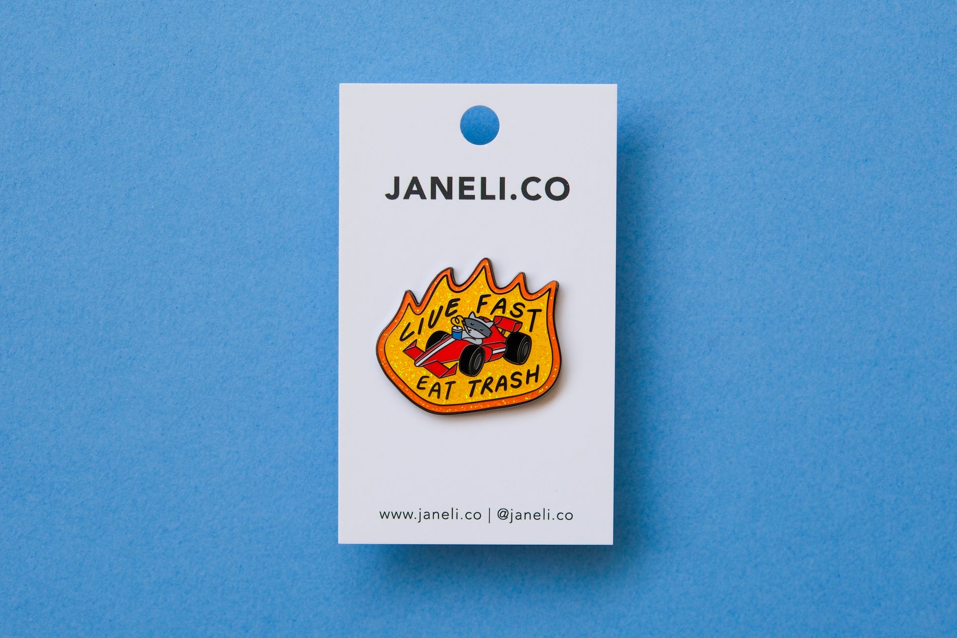 An enamel pin of a raccoon driving a racecar while sipping a soda that says "Live Fast Eat Trash" on a white JaneLi.Co backing card over a light blue background.