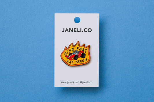 An enamel pin of a raccoon driving a racecar while sipping a soda that says "Live Fast Eat Trash" on a white JaneLi.Co backing card over a light blue background.