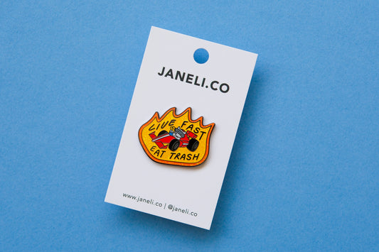 An enamel pin of a raccoon driving a racecar while sipping a soda that says "Live Fast Eat Trash" on a white JaneLi.Co backing card over a light blue background.