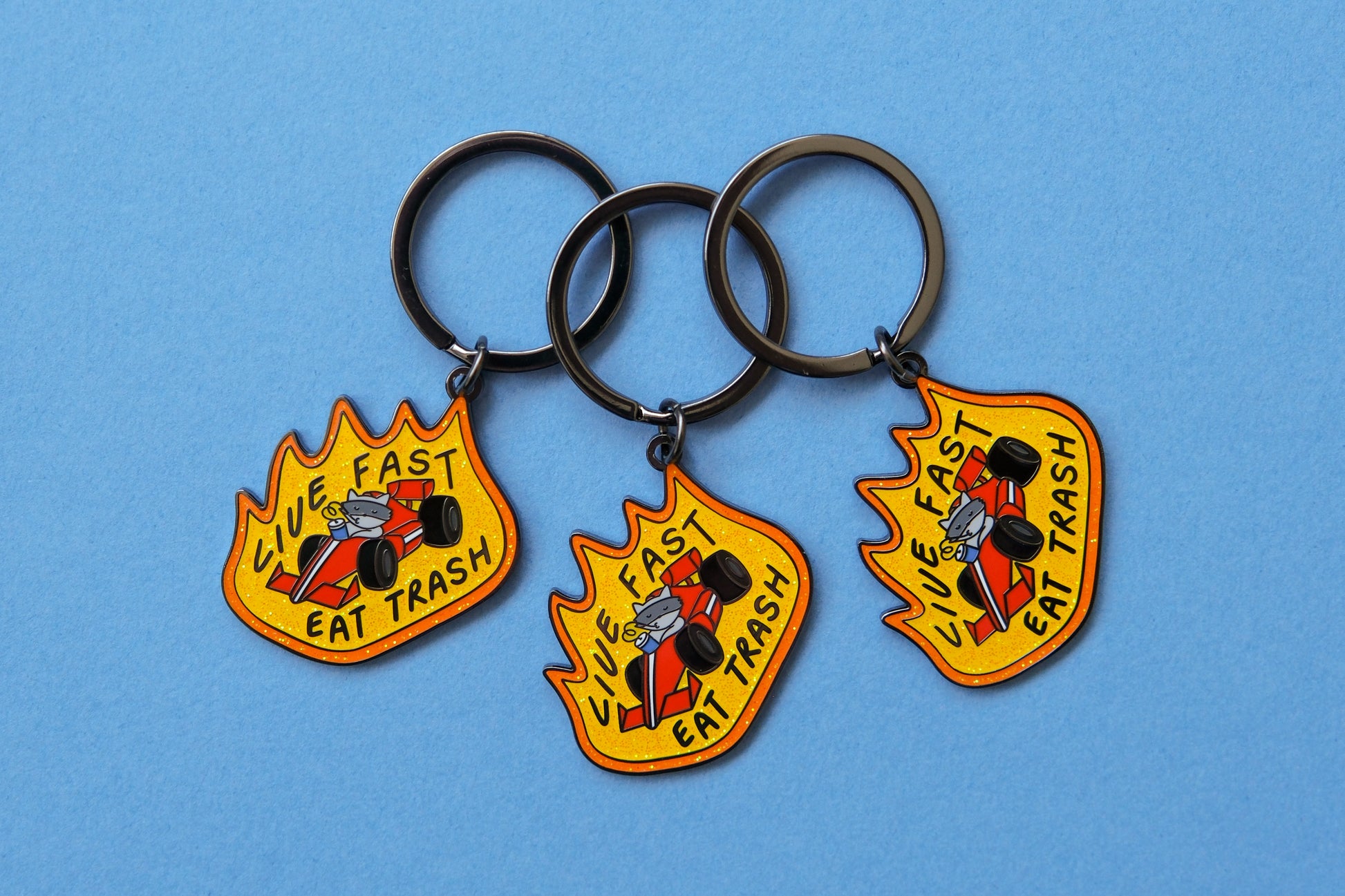 3 enamel keychains of raccoons driving a racecar while sipping a soda that says "Live Fast Eat Trash" over a light blue background.