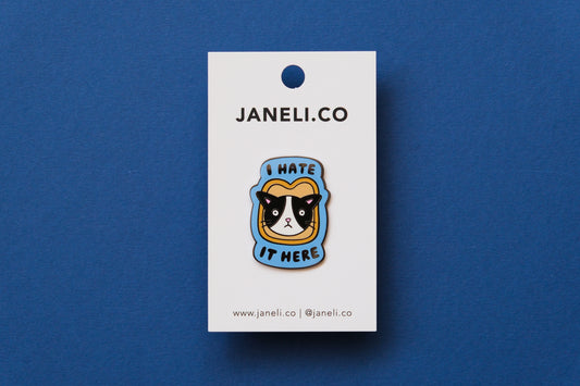 An enamel pin of a cat stuck in a piece of bread that says "I  Hate It Here" on a white JaneLi.Co backing card over a blue background.