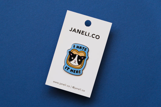 An enamel pin of a cat stuck in a piece of bread that says "I  Hate It Here" on a white JaneLi.Co backing card over a blue background.