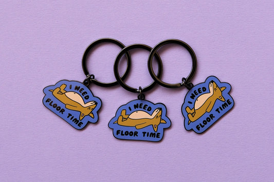 3 enamel keychains of dogs lying down that say "I Need Floor Time" over a lavender background.