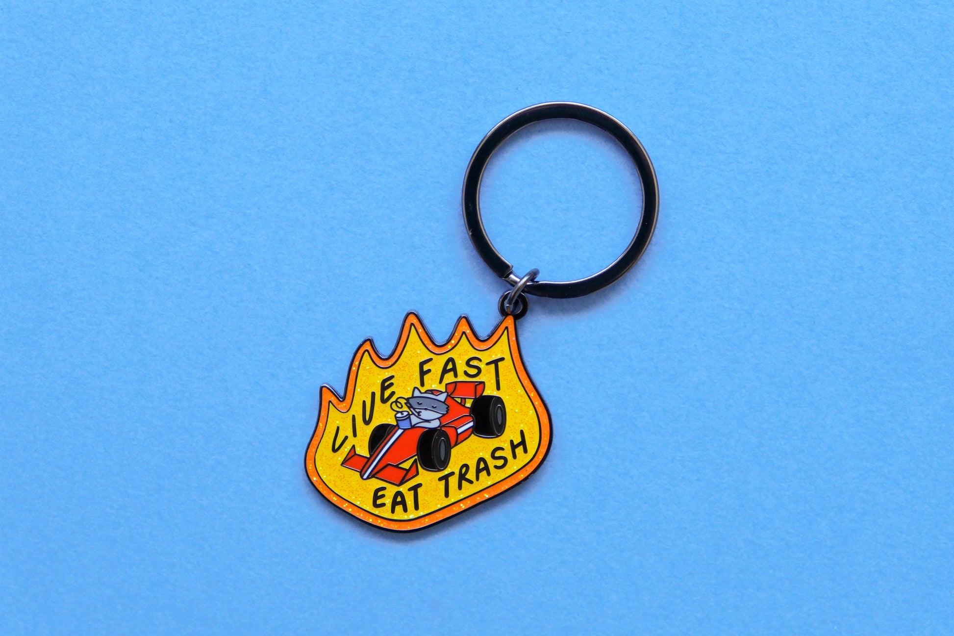An enamel keychain of a raccoon driving a racecar while sipping a soda that says "Live Fast Eat Trash" over a light blue background.