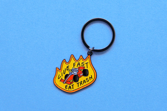 An enamel keychain of a raccoon driving a racecar while sipping a soda that says "Live Fast Eat Trash" over a light blue background.