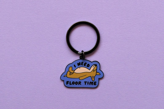 An enamel keychain of a dog lying down that says "I Need Floor Time" over a lavender background.