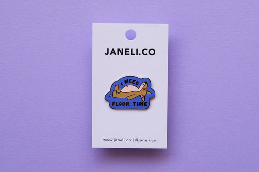 An enamel pin of a dog lying down that says "I Need Floor Time" on a white JaneLi.Co backing card over a lavender background.