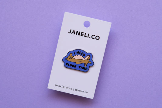 An enamel pin of a dog lying down that says "I Need Floor Time" on a white JaneLi.Co backing card over a lavender background.