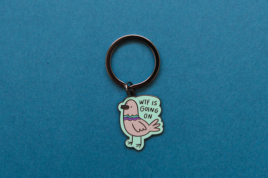An enamel keychain of a pigeon that says "WTF Is Going On" over a teal background.