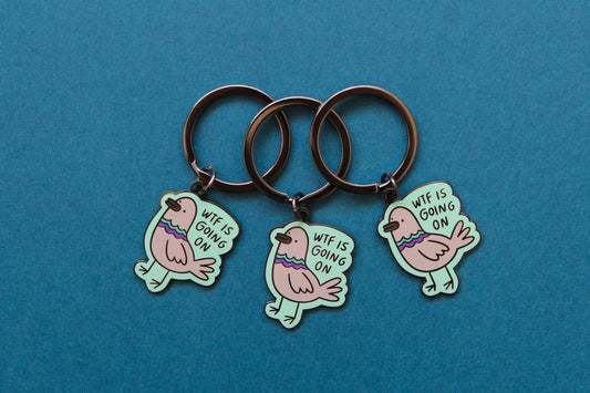 3 enamel keychains of pigeons that say "WTF Is Going On" over a teal background.