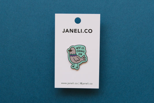An enamel pin of a pigeon that says "WTF Is Going On" on a white JaneLi.Co backing card over a teal background.