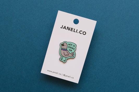 An enamel pin of a pigeon that says "WTF Is Going On" on a white JaneLi.Co backing card over a teal background.
