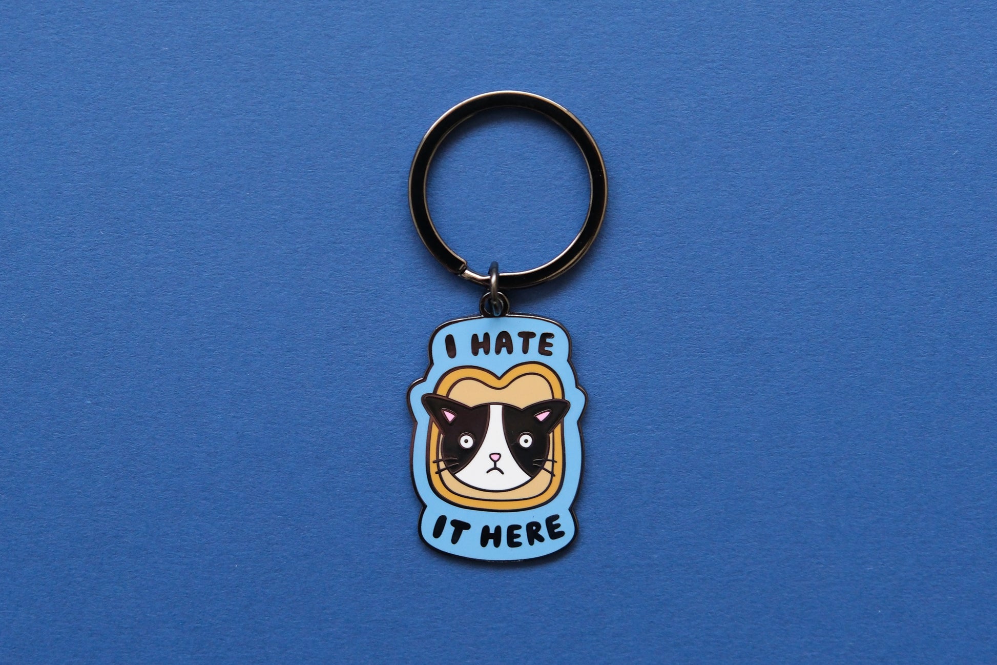 An enamel keychain of a cat stuck in a piece of bread that says "I  Hate It Here" over a blue background.