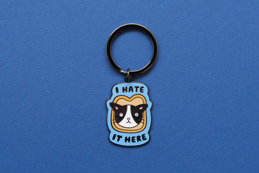 An enamel keychain of a cat stuck in a piece of bread that says "I  Hate It Here" over a blue background.