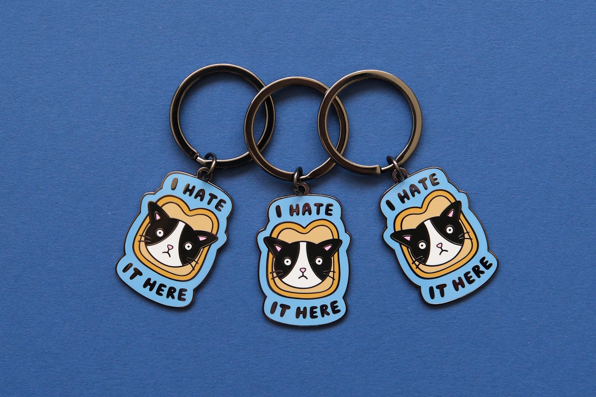 3 enamel keychains of cats stuck in a piece of bread that say "I  Hate It Here" over a blue background.