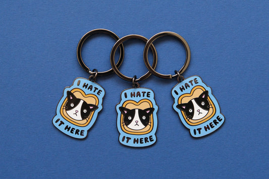 3 enamel keychains of cats stuck in a piece of bread that say "I  Hate It Here" over a blue background.