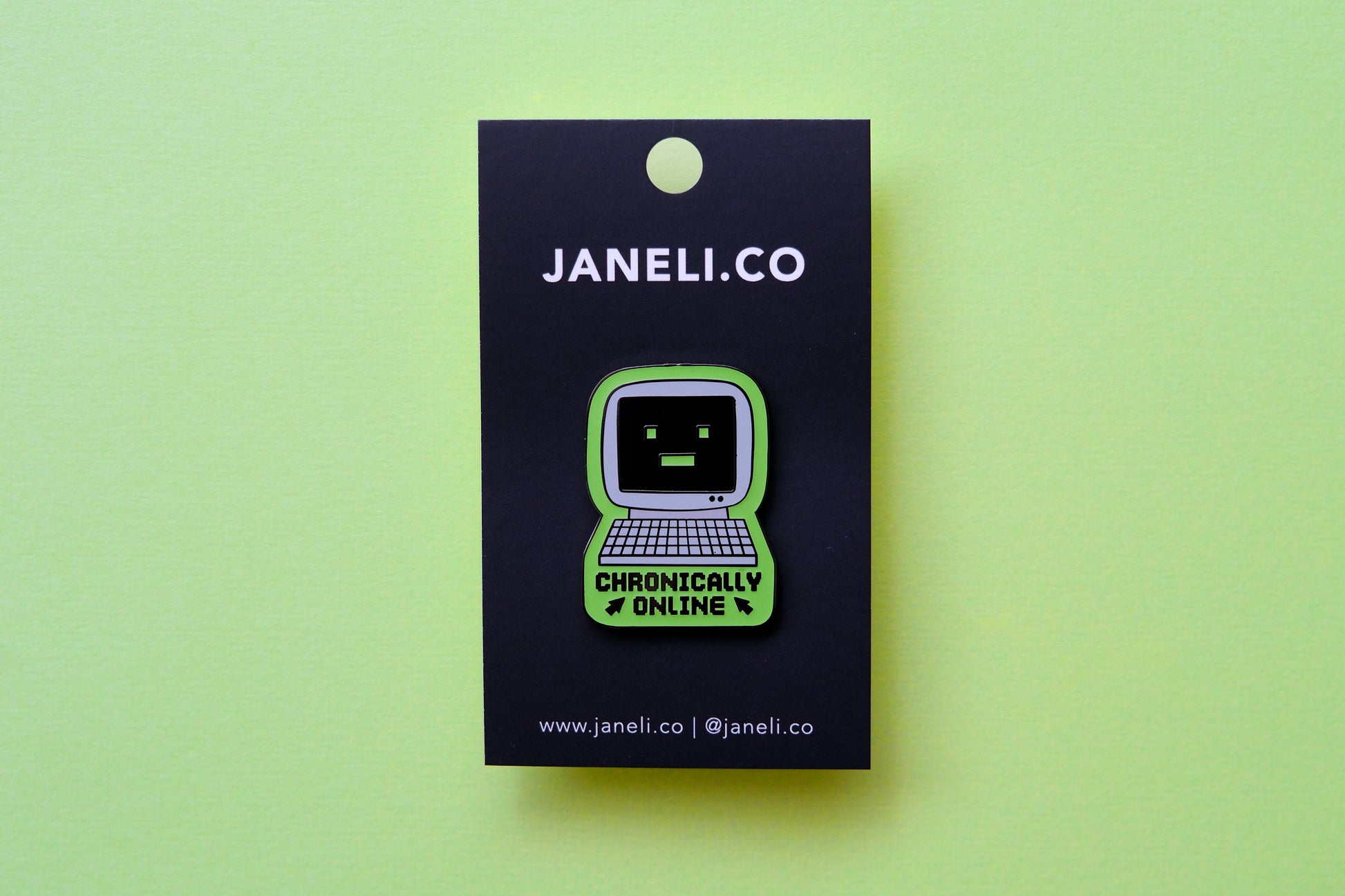 An enamel pin of a retro computer that says "Chronically Online" on a black JaneLi.Co backing card over a green background.