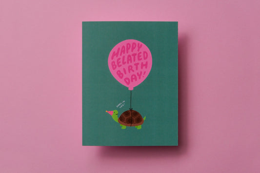 A photo of a greeting card with a turtle carrying a balloon that says "Happy belated birthday!" on a pink background.