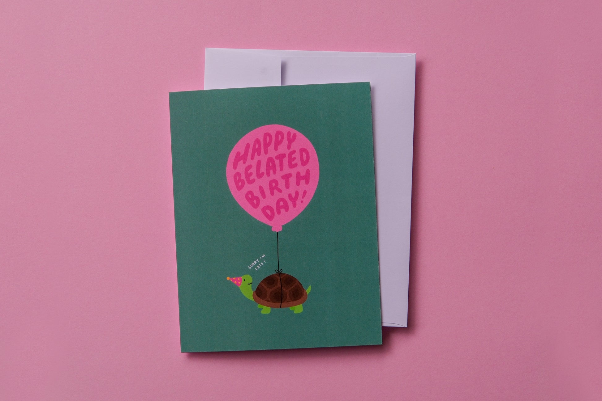 A photo of a greeting card with a turtle carrying a balloon that says "Happy belated birthday!" and a white envelope on a pink background.