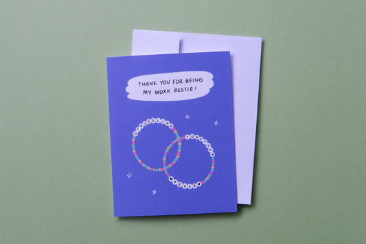 A photo of a greeting card that says "Thank you for being my work bestie!" with two friendship bracelets and a white envelope on a light green background.