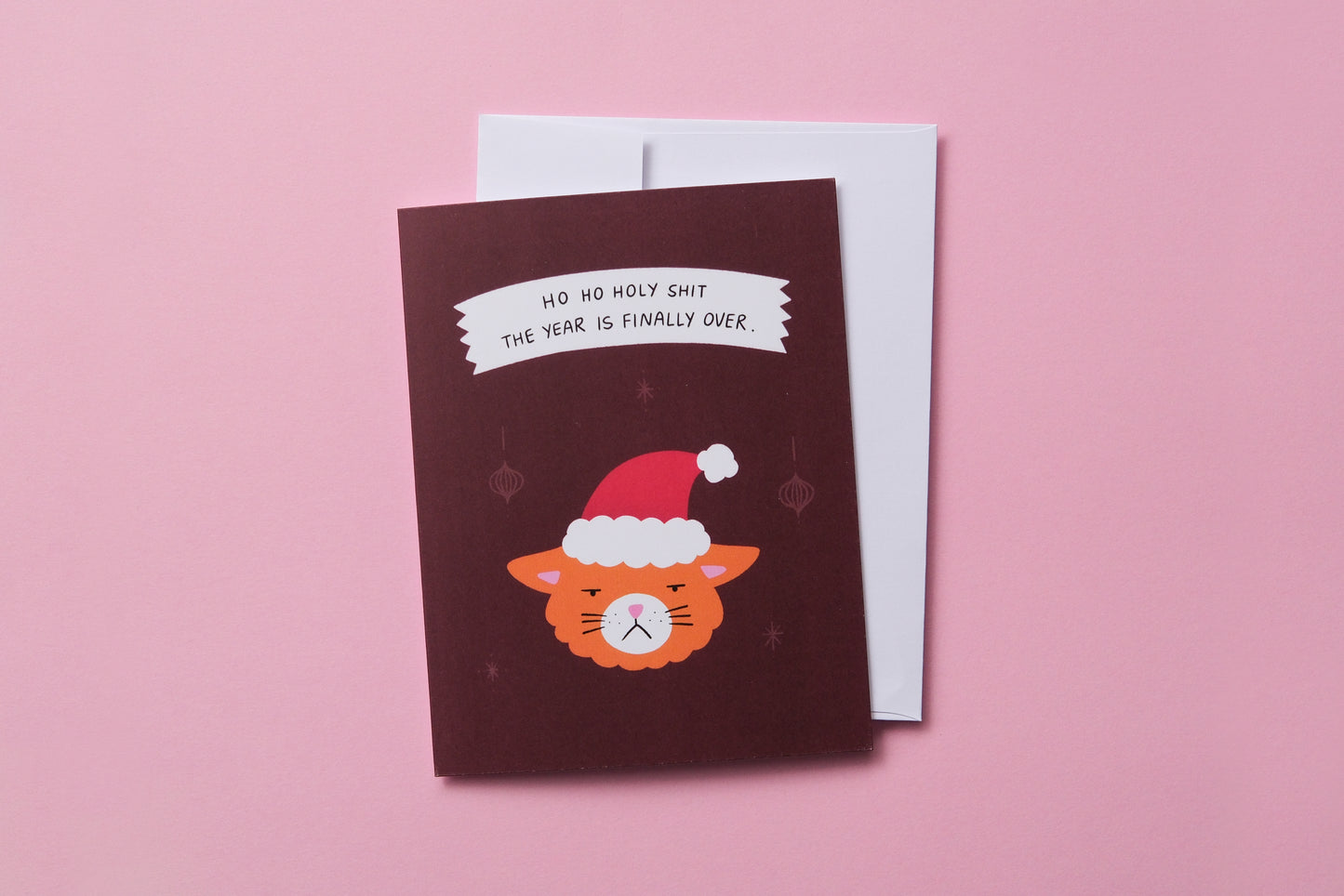 A photo of a greeting card with grumpy santa cat that says "Ho ho holy shit the year is finally over" and a white envelope on a pink background.