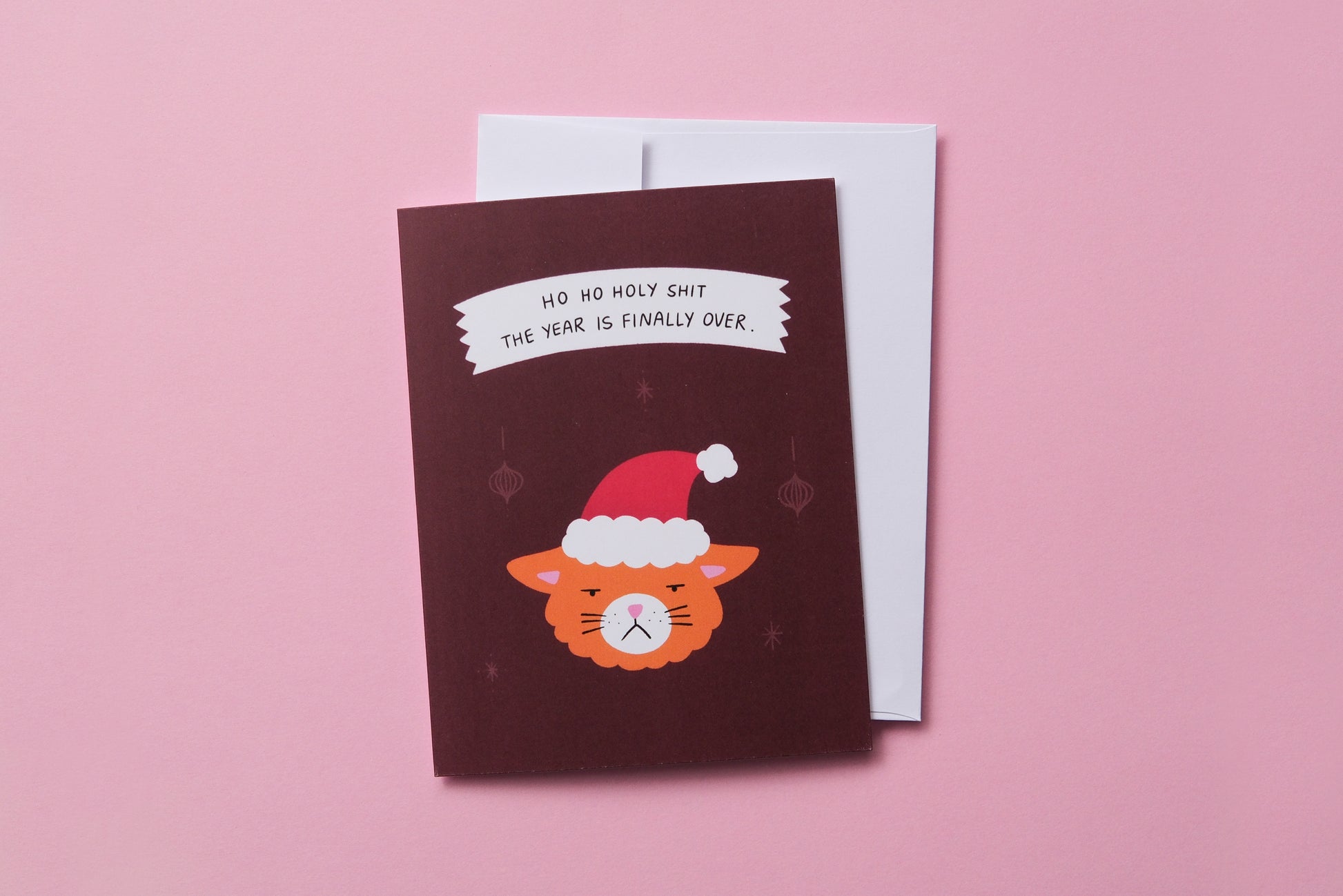 A photo of a greeting card with grumpy santa cat that says "Ho ho holy shit the year is finally over" and a white envelope on a pink background.