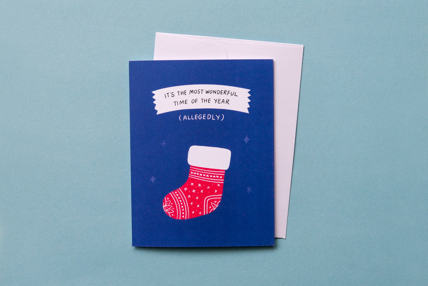 A photo of a greeting card with a stocking that says "It's the most wonderful time of the year (allegedly)" and a white envelope on a blue background.