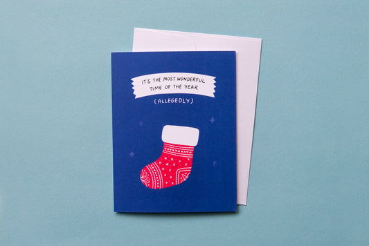 A photo of a greeting card with a stocking that says "It's the most wonderful time of the year (allegedly)" and a white envelope on a blue background.